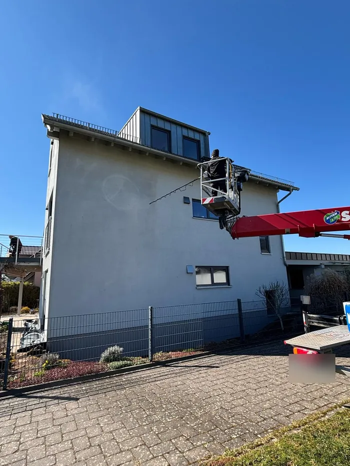 Hebebühne vor einer grauen Putzfassade, ein Mitarbeiter reinigt die Wand von oben